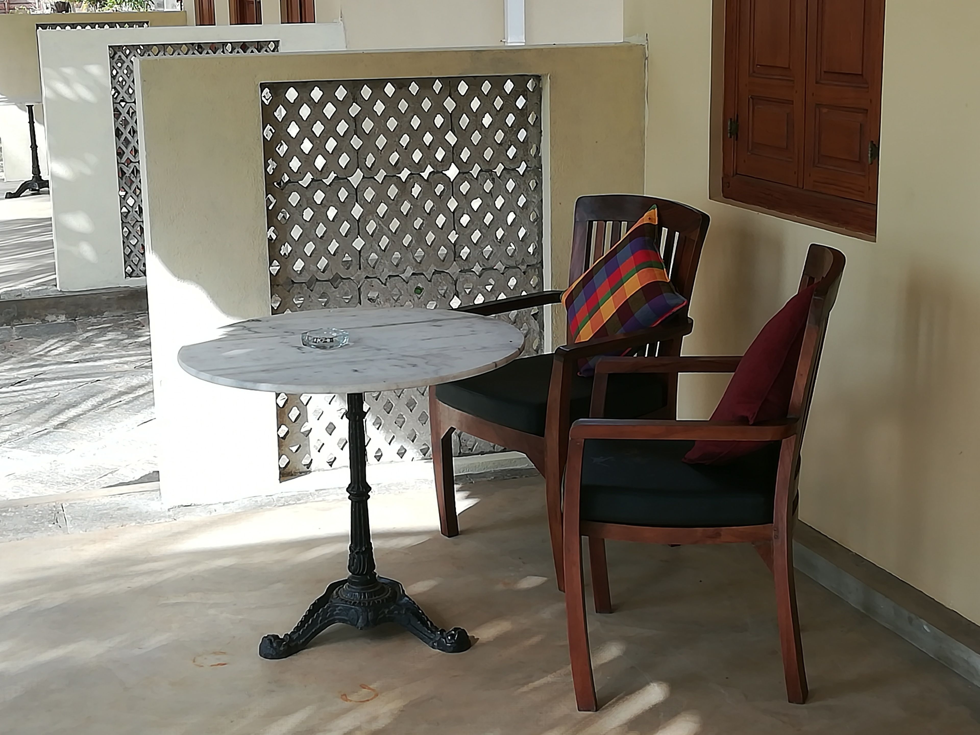 Une table ronde avec un pied en métal, une chaise en bois avec un coussin coloré, et un mur à motifs en arrière-plan.