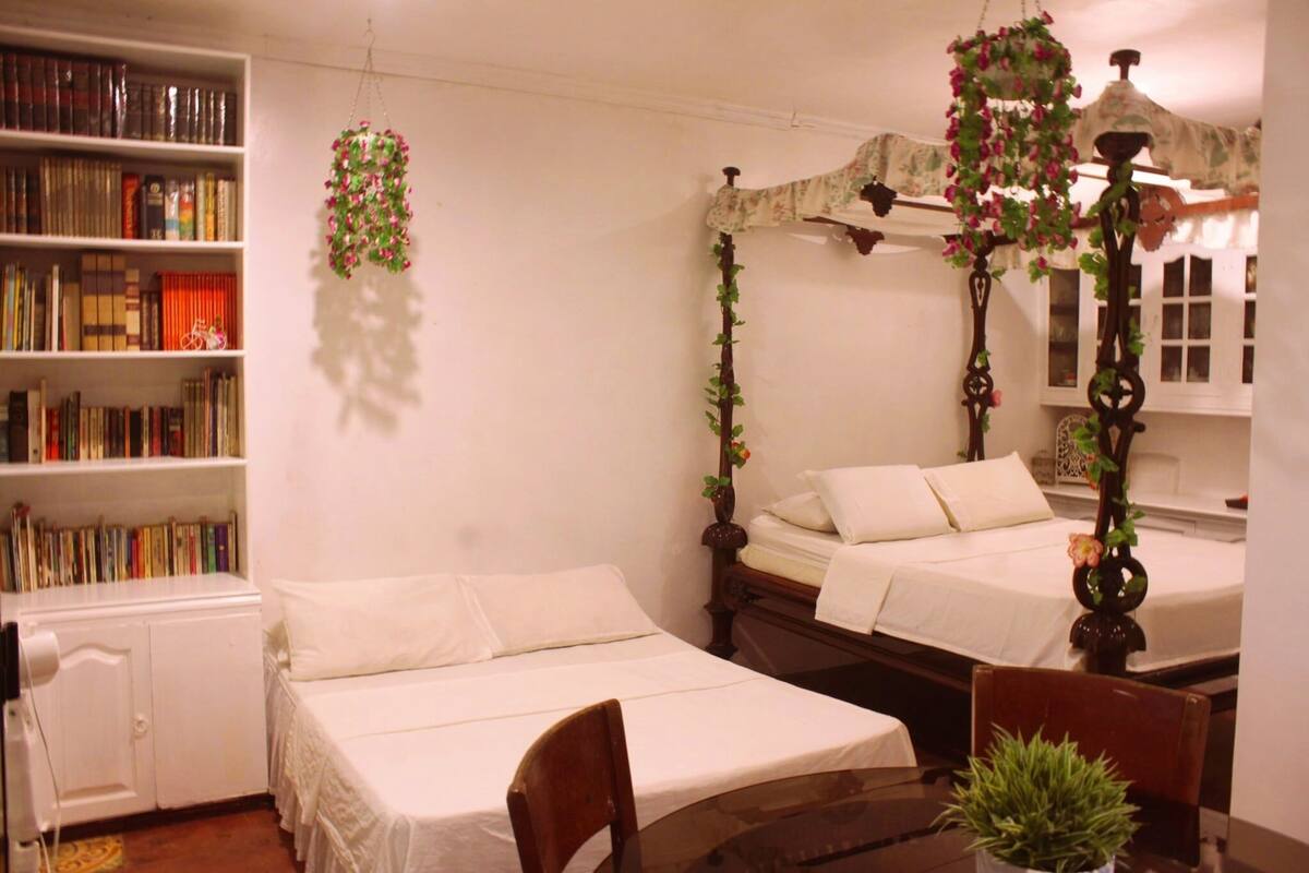 A room with two single beds, a wooden nightstand, a small table with a plant, a bookshelf filled with books, and a hanging floral decoration.