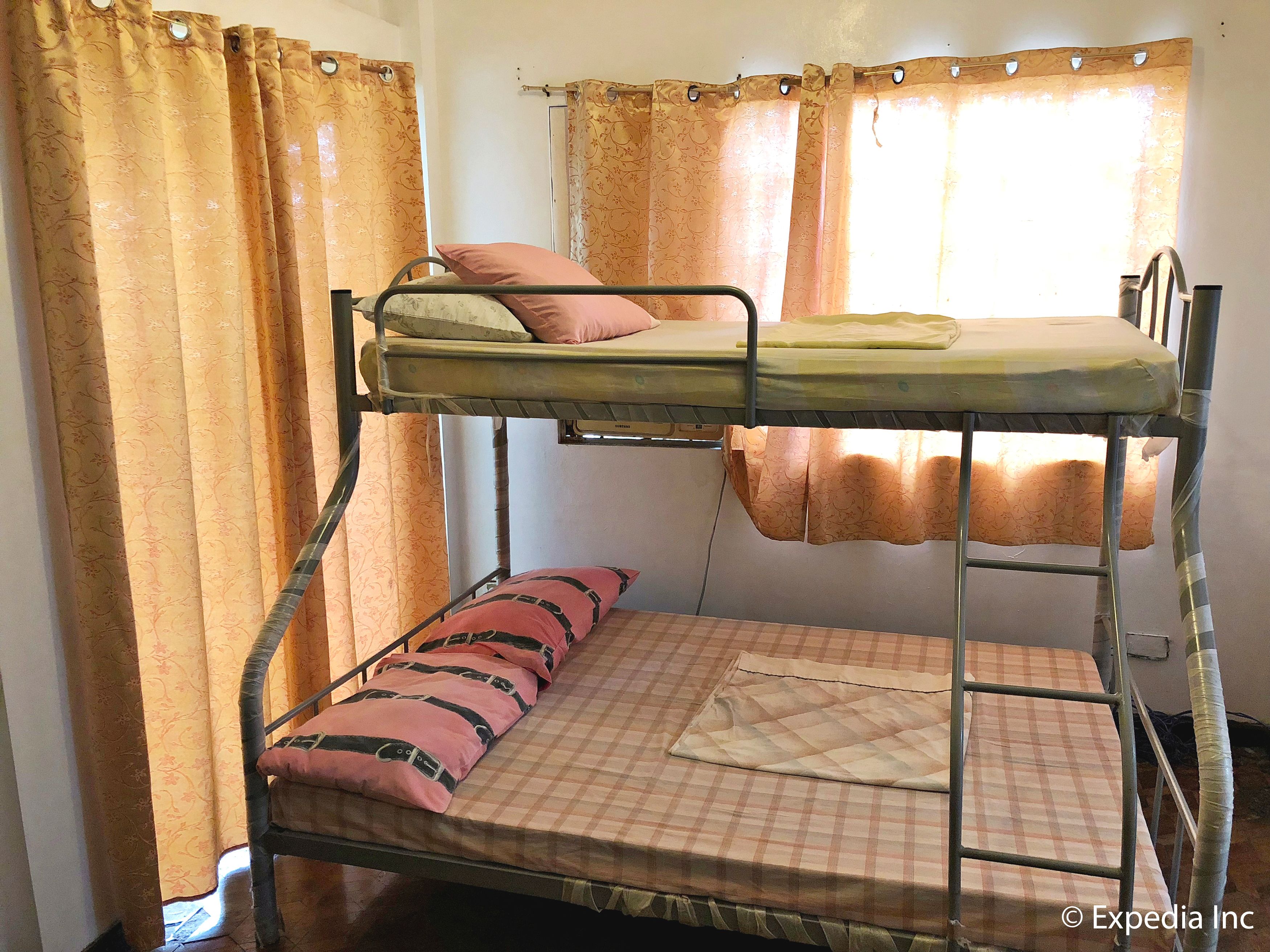 Una cama litera con colchón a cuadros y almohadas rosas, una ventana con cortinas y estructura metálica.