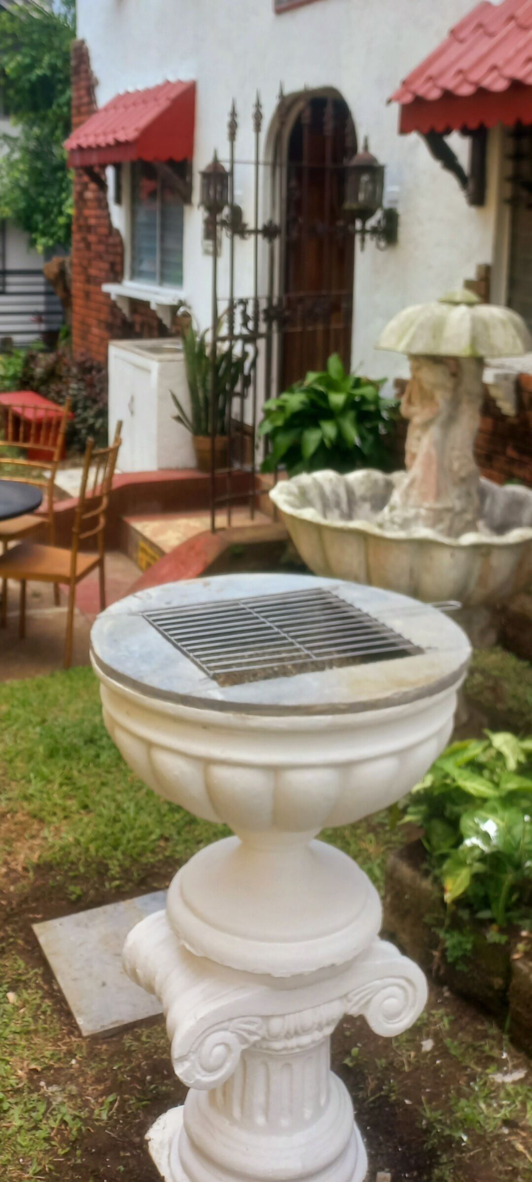 A white outdoor fountain with a decorative pedestal, surrounded by greenery and a brick wall.