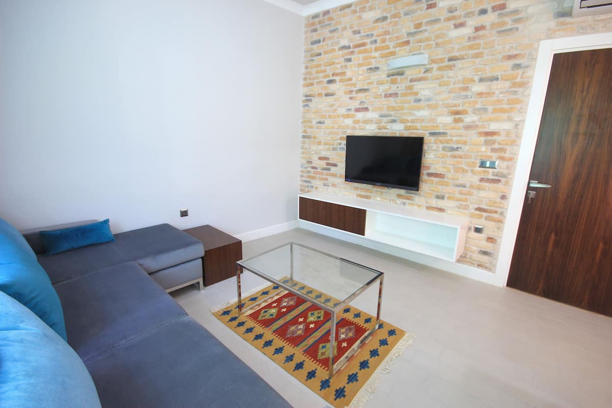 Ein modernes Wohnzimmer mit Ecksofa, Glastisch, Flachbildfernseher an einer Ziegelwand und Holztür.