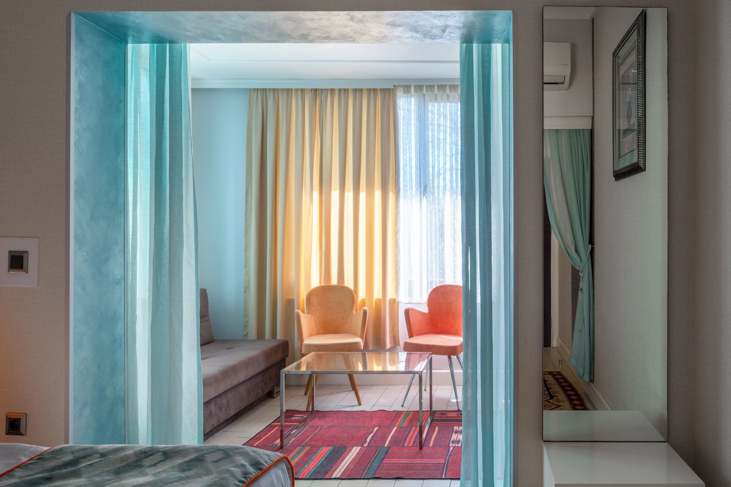 Une chambre d’hôtel comprenant un lit, un canapé, deux fauteuils, une table basse en verre et une vue sur un balcon avec des rideaux.