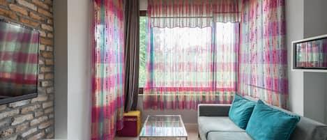 A living room with a sofa, a coffee table, a TV mounted on a brick wall, and colorful curtains.