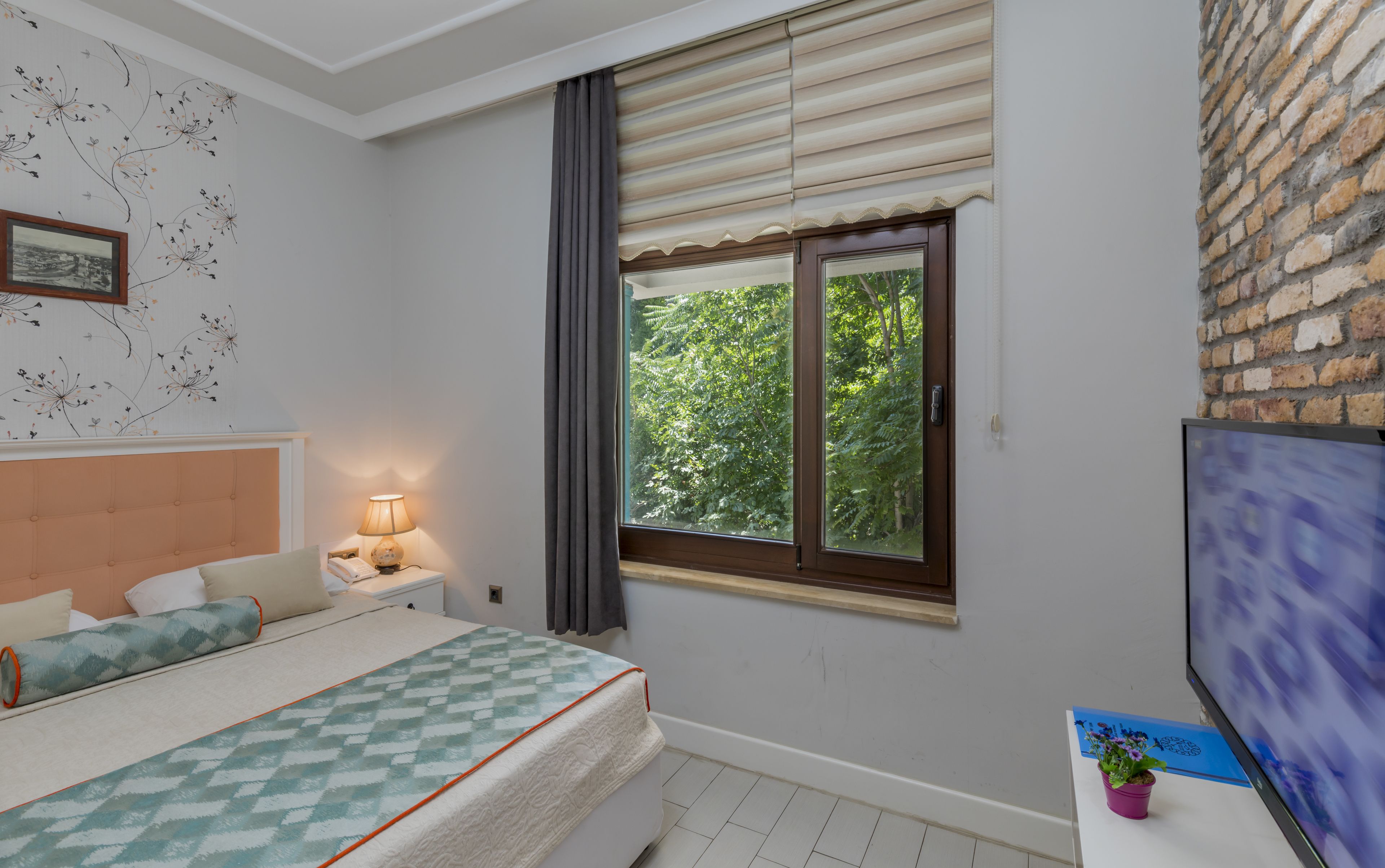 Ein Schlafzimmer mit Bett, Nachttisch mit Lampe, einem Fenster mit Jalousien, einem Fernseher und einer Ziegelwand.