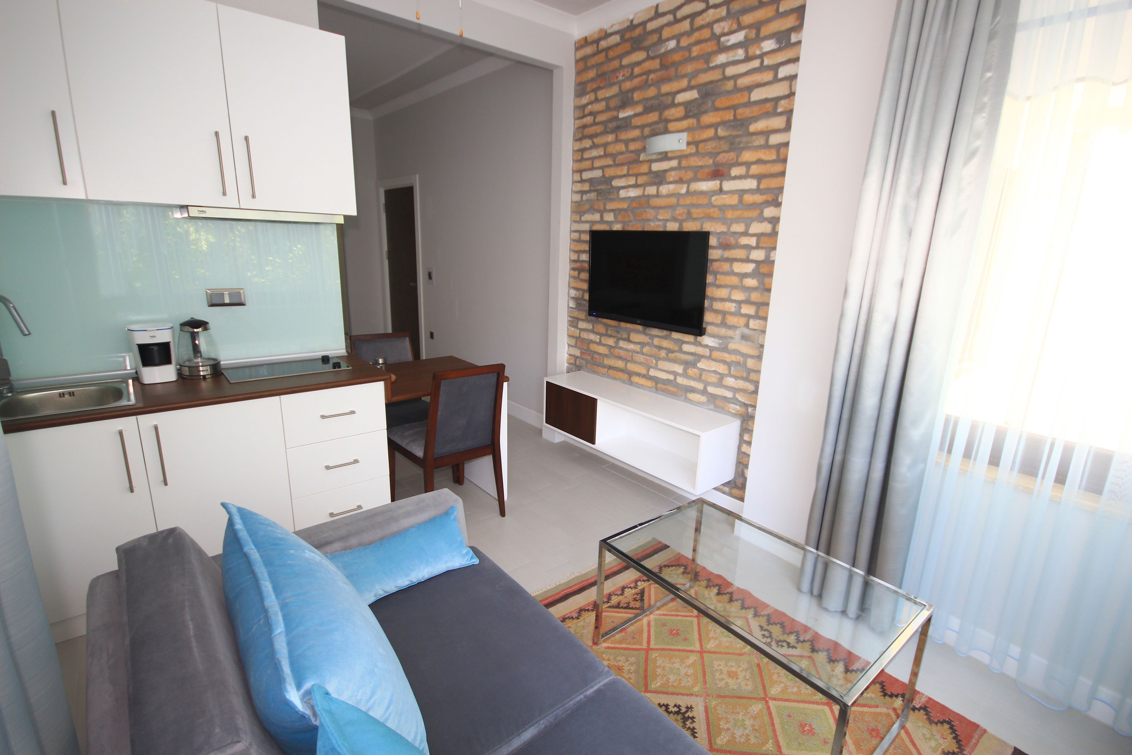 A modern living room with a sofa, a glass coffee table, a TV mounted on a brick wall, and a kitchen area with cabinets and a coffee machine.