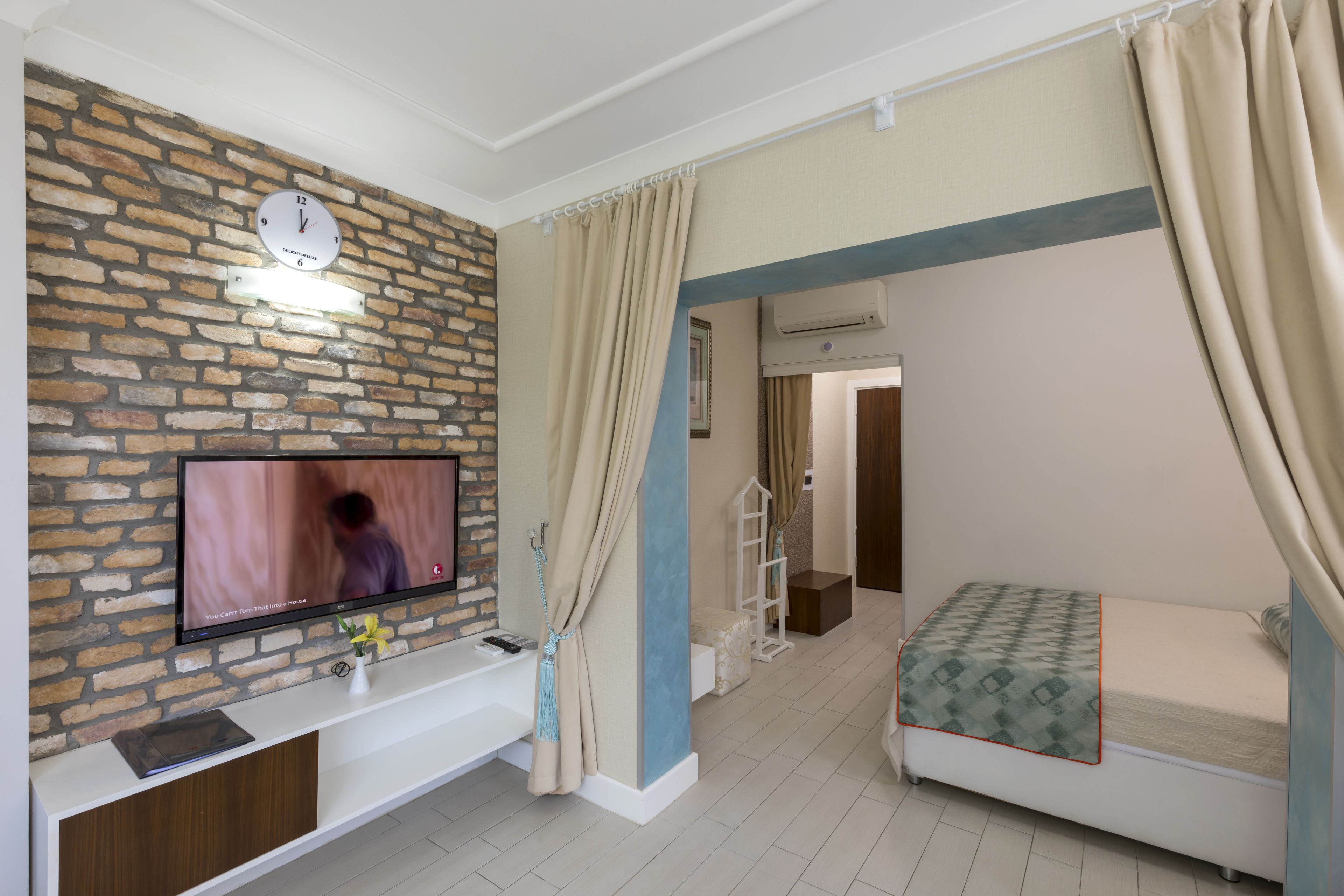 Un dormitorio con pared de ladrillo, un televisor sobre una mesa consola, una cama con colcha estampada y una mesita de noche con un florero.