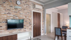 A living room with a brick wall, a flat-screen TV, a glass coffee table, and a dining area with a table and chairs.