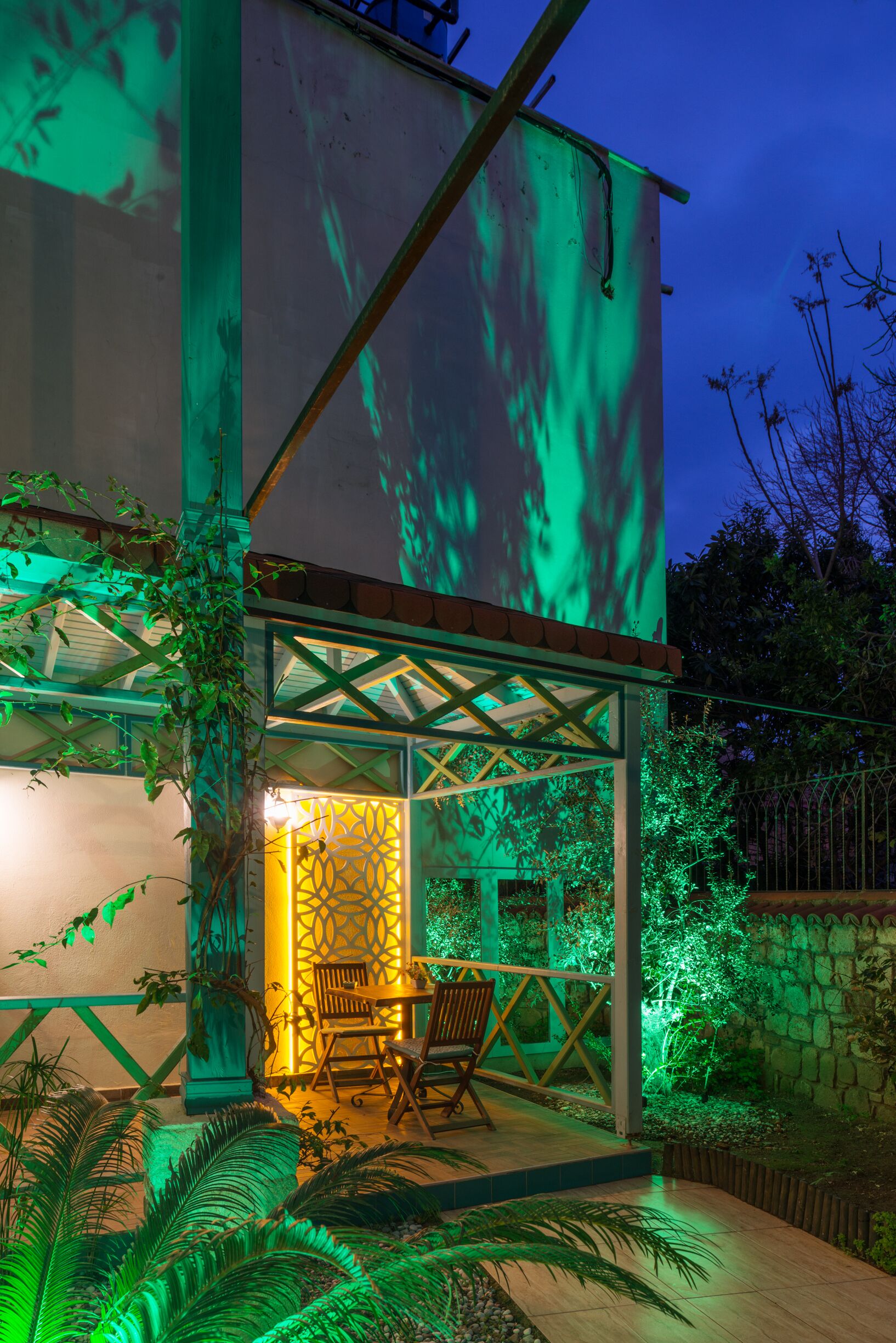 A modern house with a glass facade, outdoor seating area, and green ambient lighting.