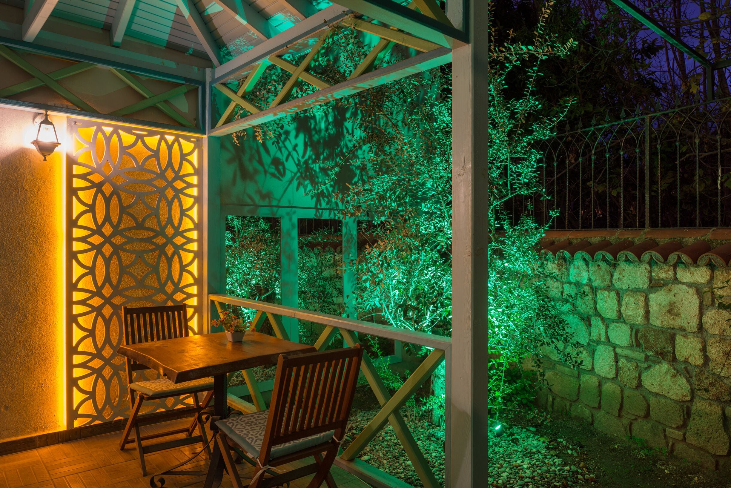 A covered outdoor seating area with a table and chairs, surrounded by a stone wall and greenery.