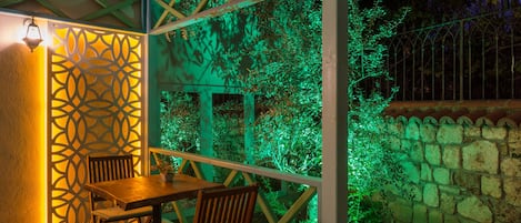 A covered outdoor seating area with a table and chairs, surrounded by a stone wall and greenery.