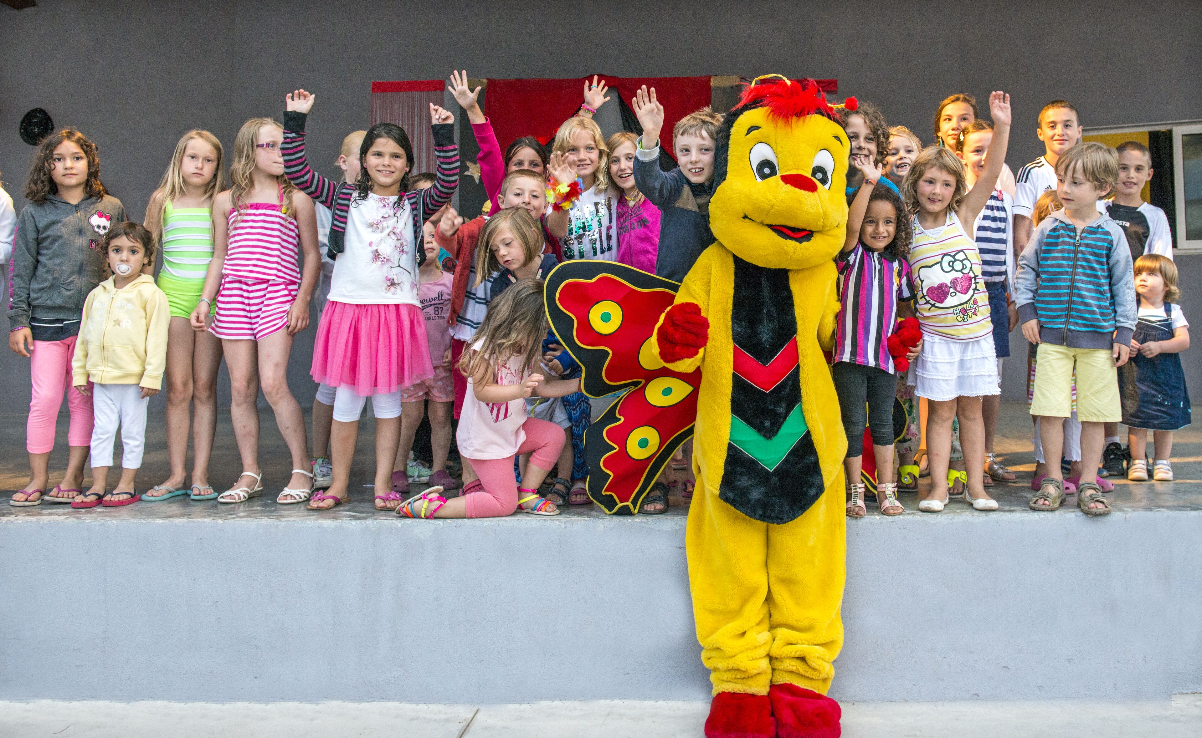 A mascot in a yellow costume with red accents standing in front of a group of children and adults.