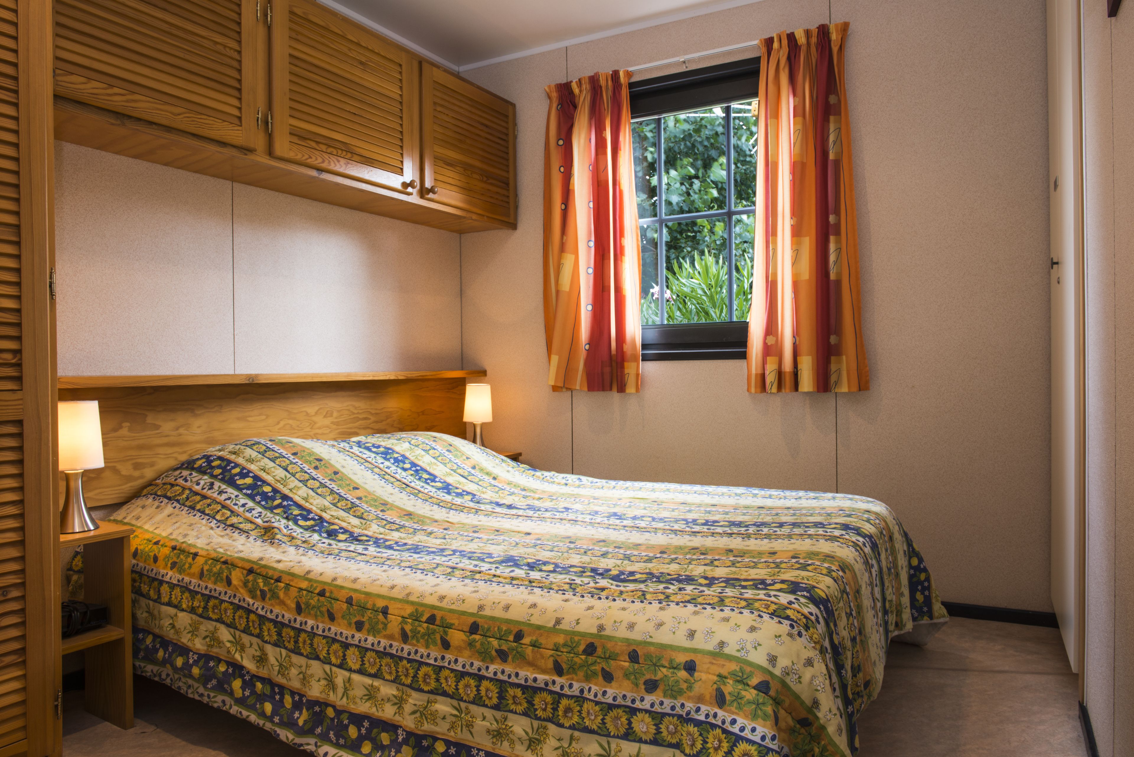 Een slaapkamer met een bed, houten kasten, een raam met gordijnen en een nachtkastje met een lamp.
