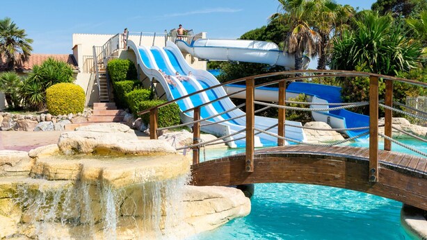 A water park with a bridge over a pool, slides, and palm trees. - Les Mûriers (Vendres)