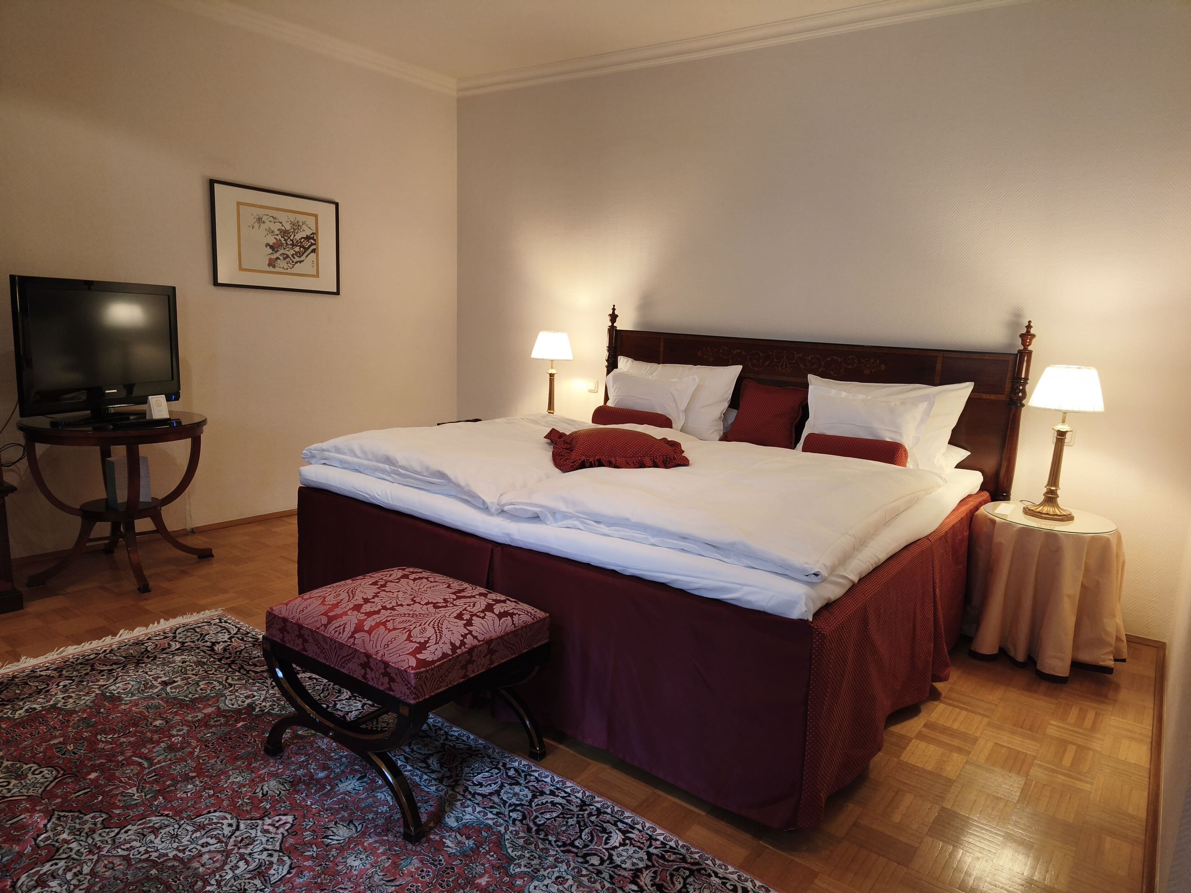 A neatly made bed with white and red bedding, two bedside lamps, a television on a small table, and a patterned rug.