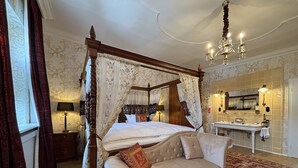 Un lit à baldaquin à quatre poteaux, agrémenté d’un dais, un canapé, une table de chevet, une lampe, un lustre, un miroir et une salle de bain avec un lavabo et un porte-serviettes.