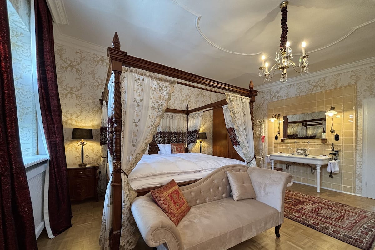 A four-poster canopy bed with a canopy, a sofa, a bedside table, a lamp, a chandelier, a mirror, and a bathroom with a sink and towel rack.