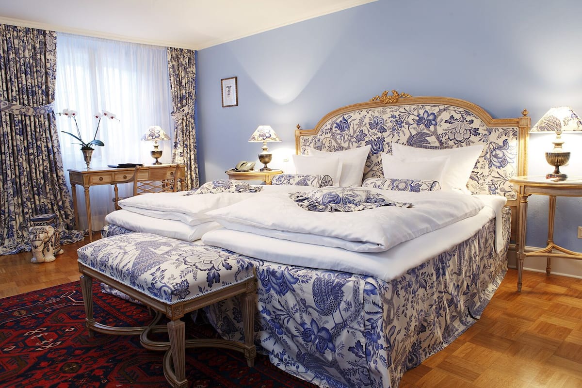 A bedroom with a large bed, two bedside tables, a nightstand, a red patterned rug, and a window with curtains.