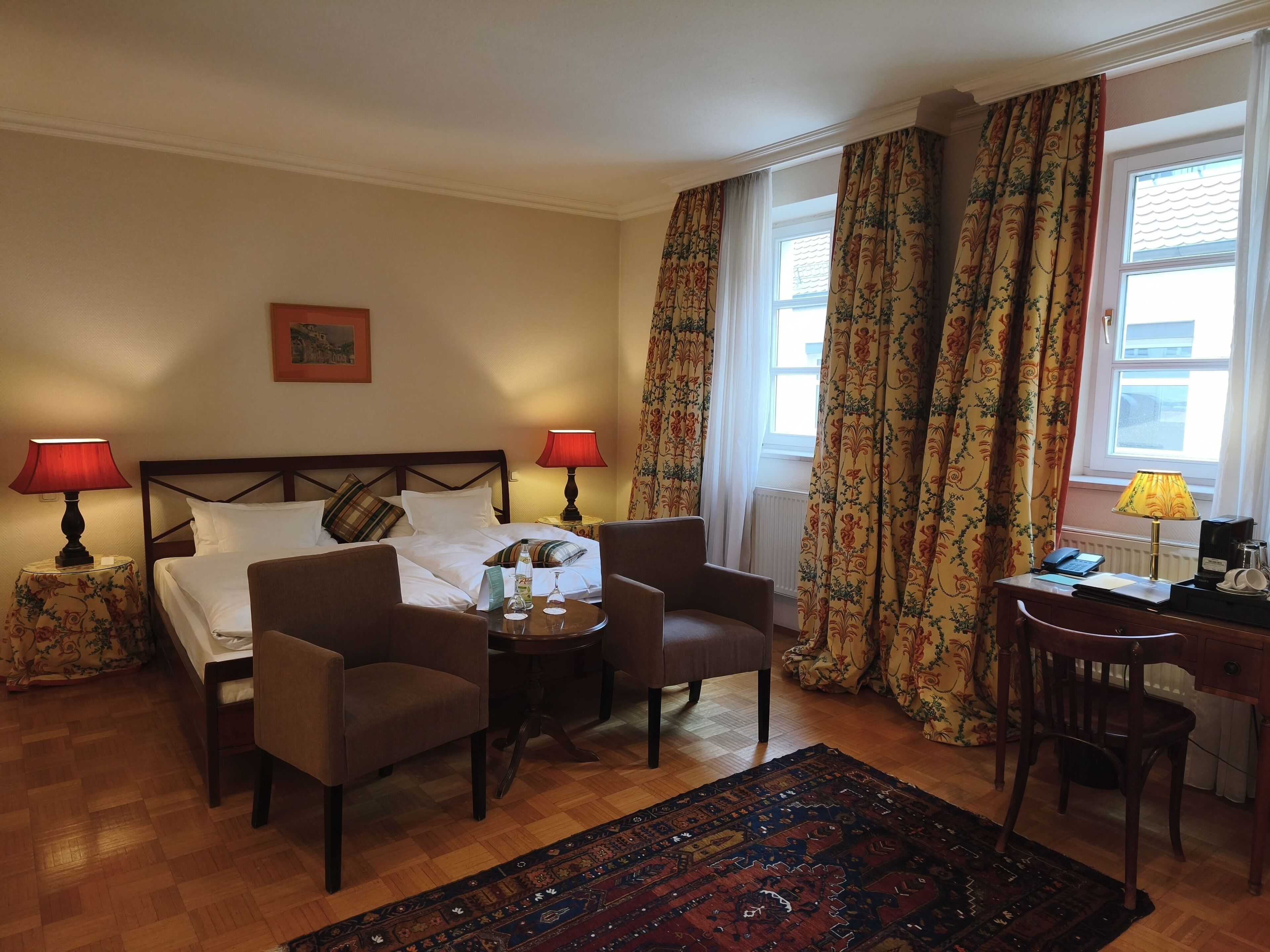 A hotel room with a bed, two chairs, a small table, a desk, and a computer. There are two lamps, a window with curtains, and a radiator.