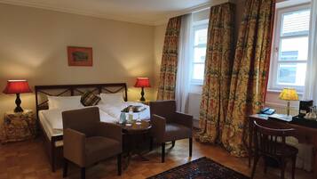 A hotel room with a bed, two chairs, a small table, a desk, and a computer. There are two lamps, a window with curtains, and a radiator.