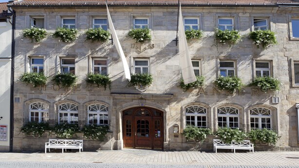 Un bâtiment historique doté d’une façade en pierre, de fenêtres en arc de cercle et d’une entrée centrale avec une porte en bois.