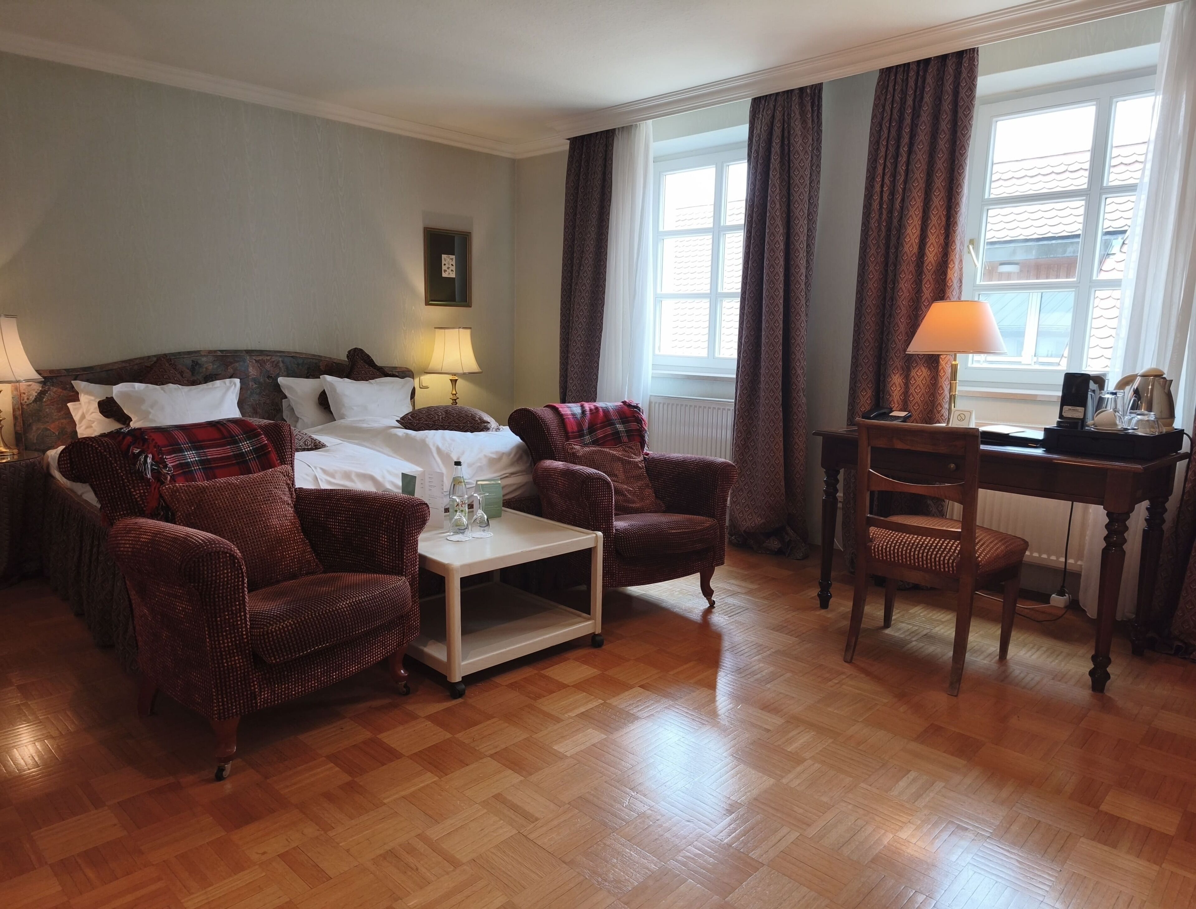 A hotel room with a bed, two armchairs, a small table, a desk, and a window with curtains.