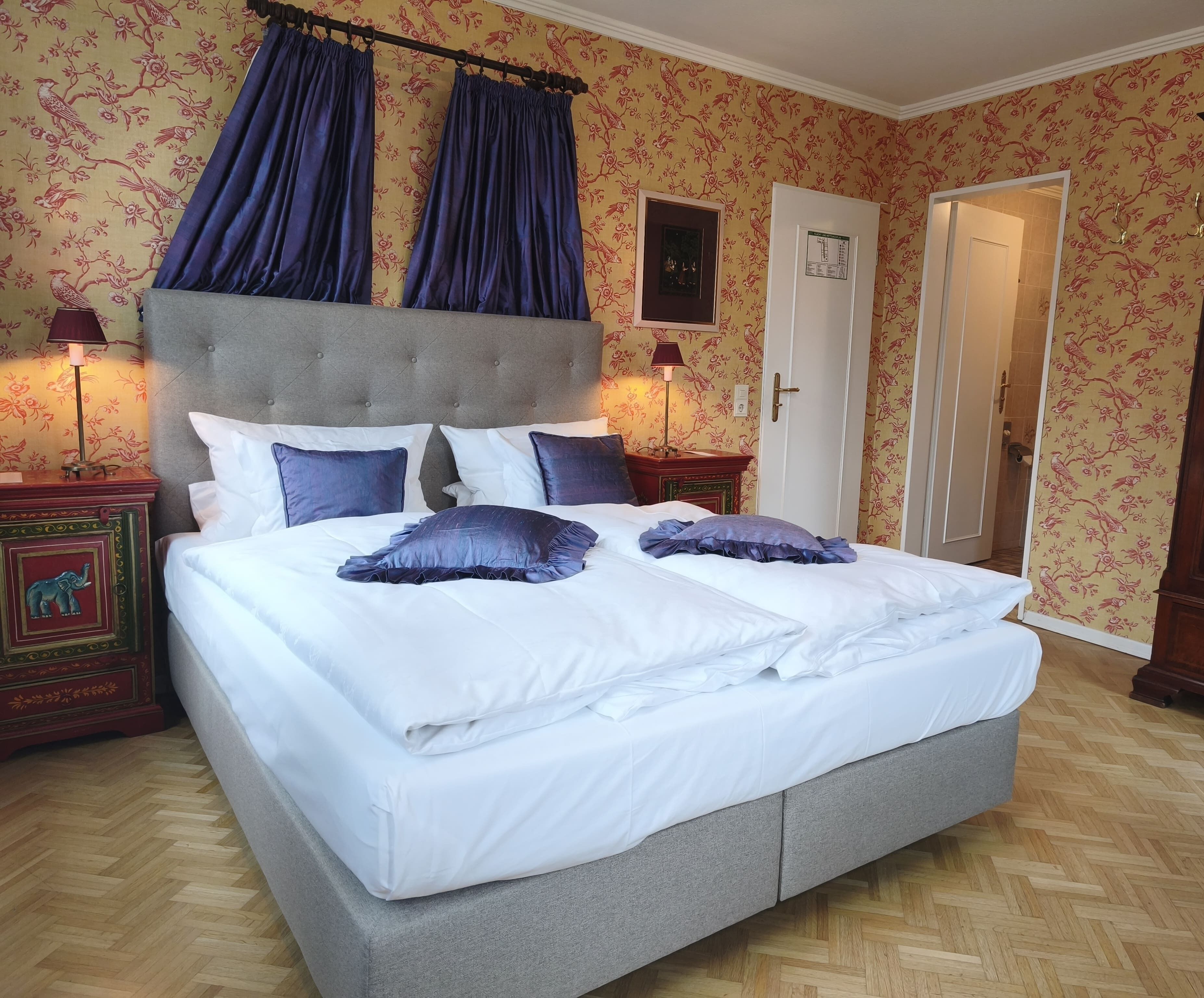 A large bed with white linens and blue pillows, a grey upholstered headboard, and a wooden floor.