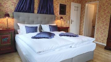 A large bed with white linens and blue pillows, a grey upholstered headboard, and a wooden floor.