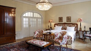 Une chambre à coucher avec un grand lit, des fauteuils à motifs floraux, une armoire en bois et un lustre.
