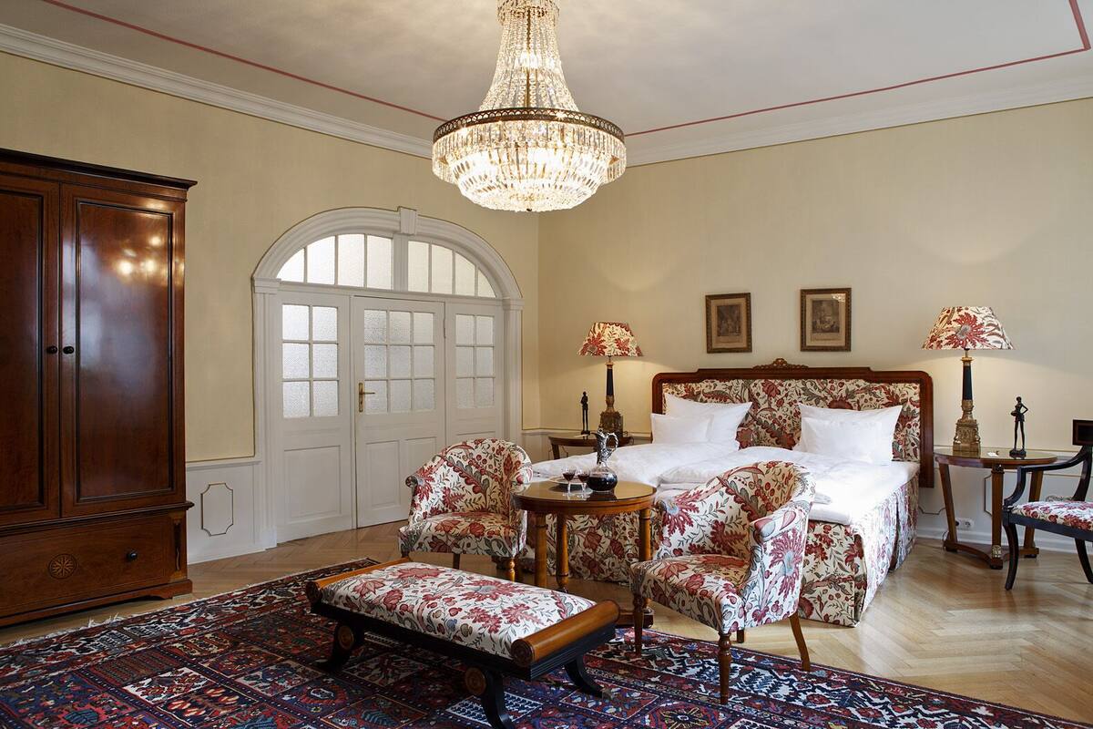 A bedroom with a large bed, floral-patterned chairs, a wooden cabinet, and a chandelier.