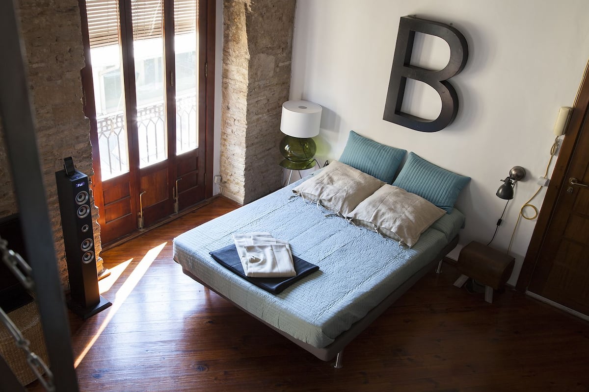 A bedroom with a bed, a lamp, a large 'B' wall decoration, and a wooden floor.