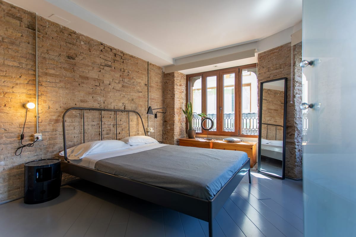 A modern bedroom with a brick wall, a metal bed frame, a bedside table with a lamp, and a view of the outside balcony.