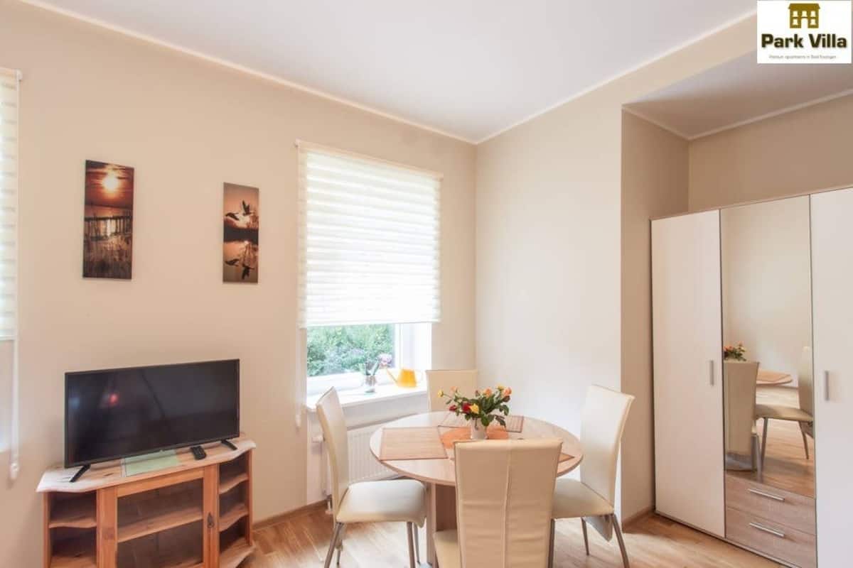 Een moderne woonkamer met een flatscreen televisie op een houten standaard, een eettafel met stoelen en een grote kledingkast.