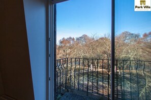 Uitzicht vanuit een raam met een metalen hekwerk, uitkijkend op een park met bomen en een gebouw op de achtergrond.