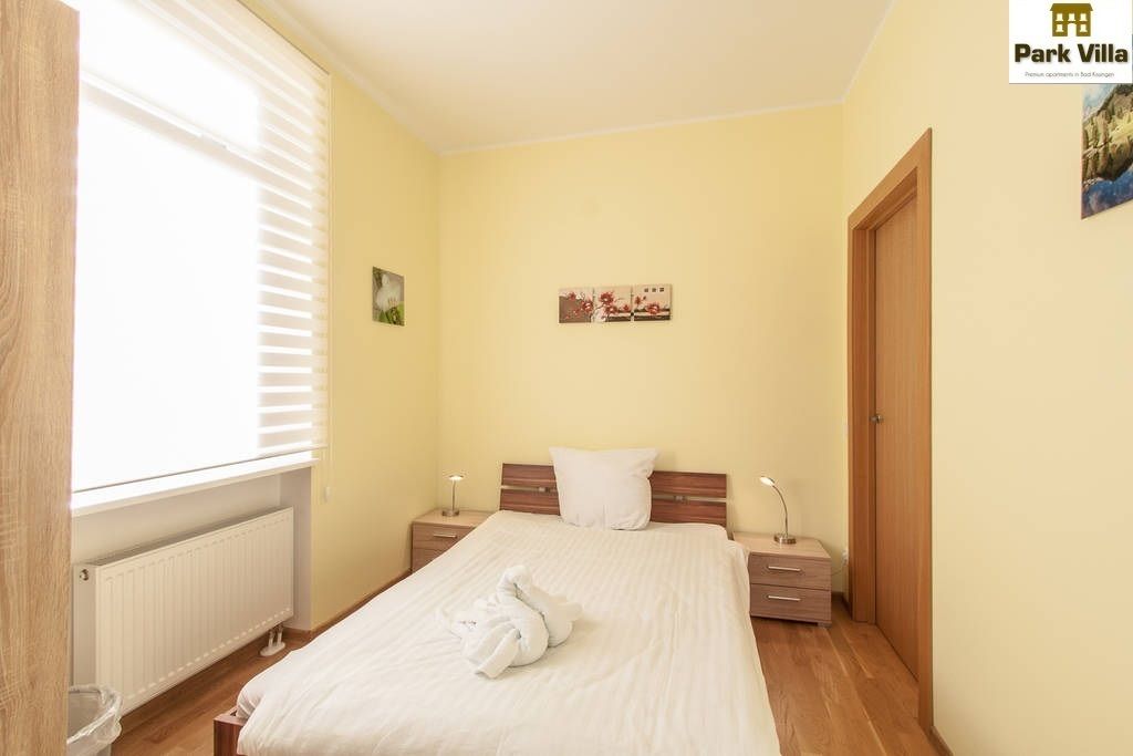 A bedroom with a bed, bedside tables, a radiator, and a window with blinds.