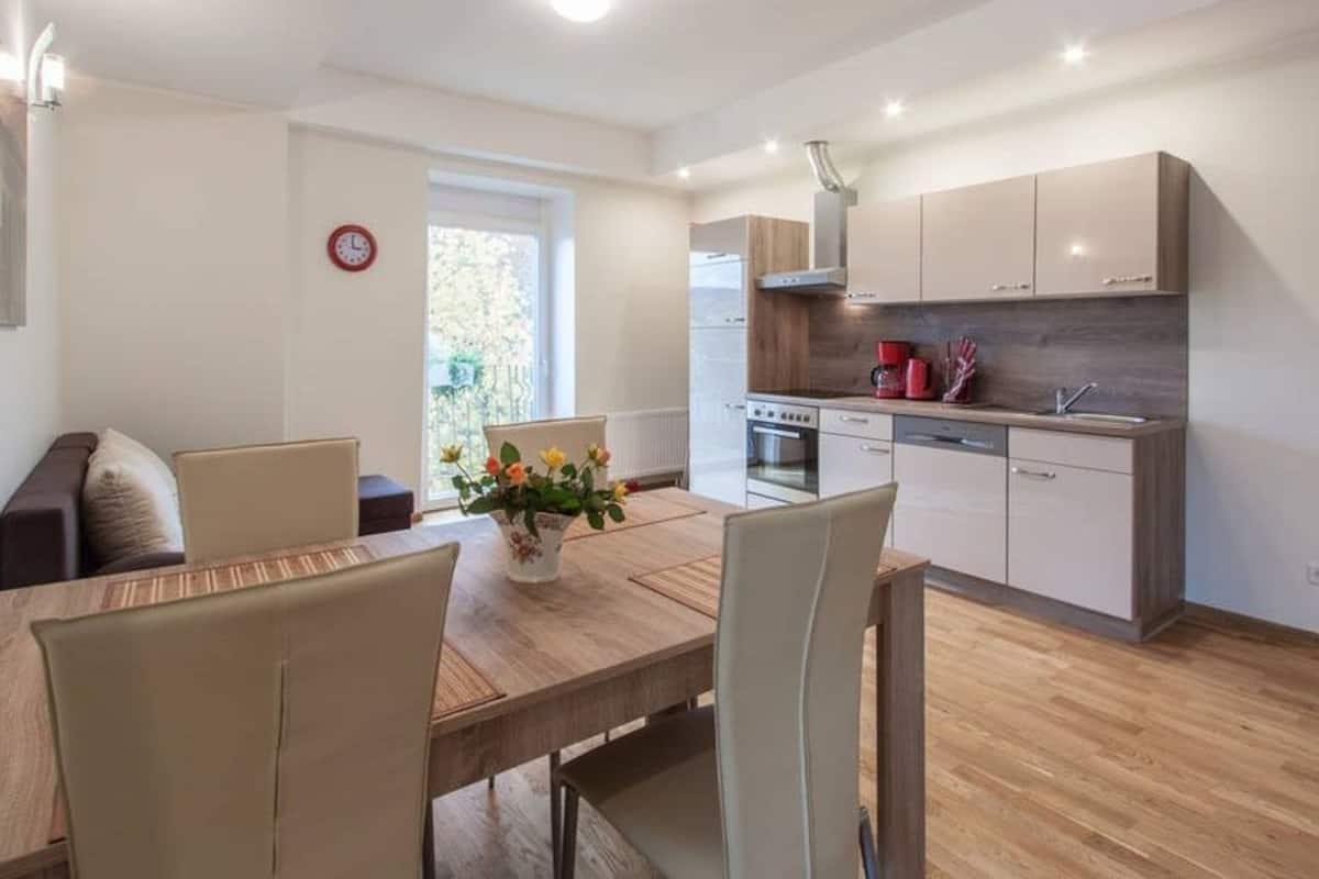 Een moderne keuken met een houten eettafel en stoelen, een inbouwoven en een raam met uitzicht op groen.