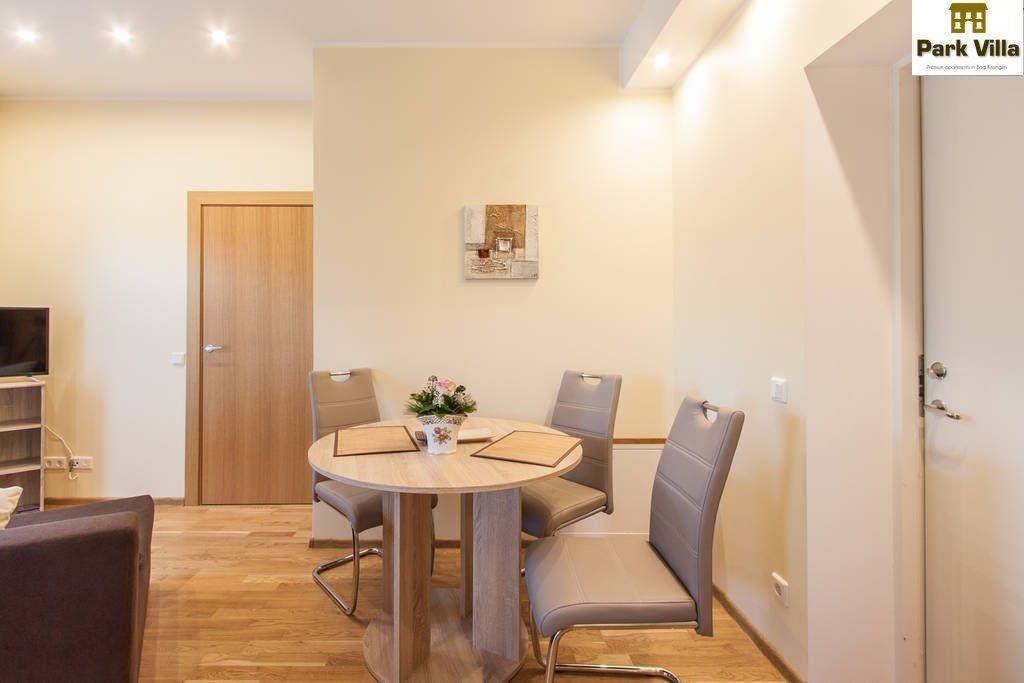 Ein modernes Wohnzimmer mit einem runden Holztisch, vier grauen Stühlen, einem Fernseher und einer Holztür.