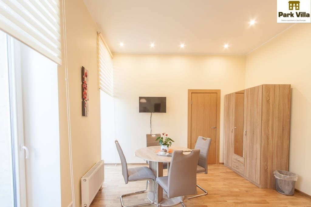 A modern living room with a round dining table, grey chairs, a TV mounted on the wall, and wooden wardrobes.