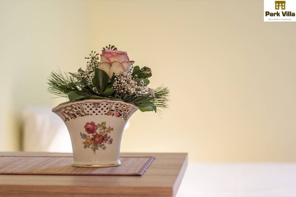A white ceramic vase with floral patterns and a pink rose on a wooden table.