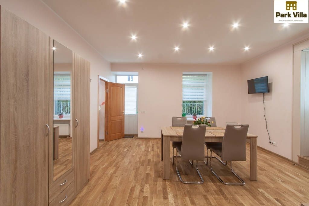 A modern dining area with a wooden table and chairs, a TV mounted on the wall, and a door leading to another room.