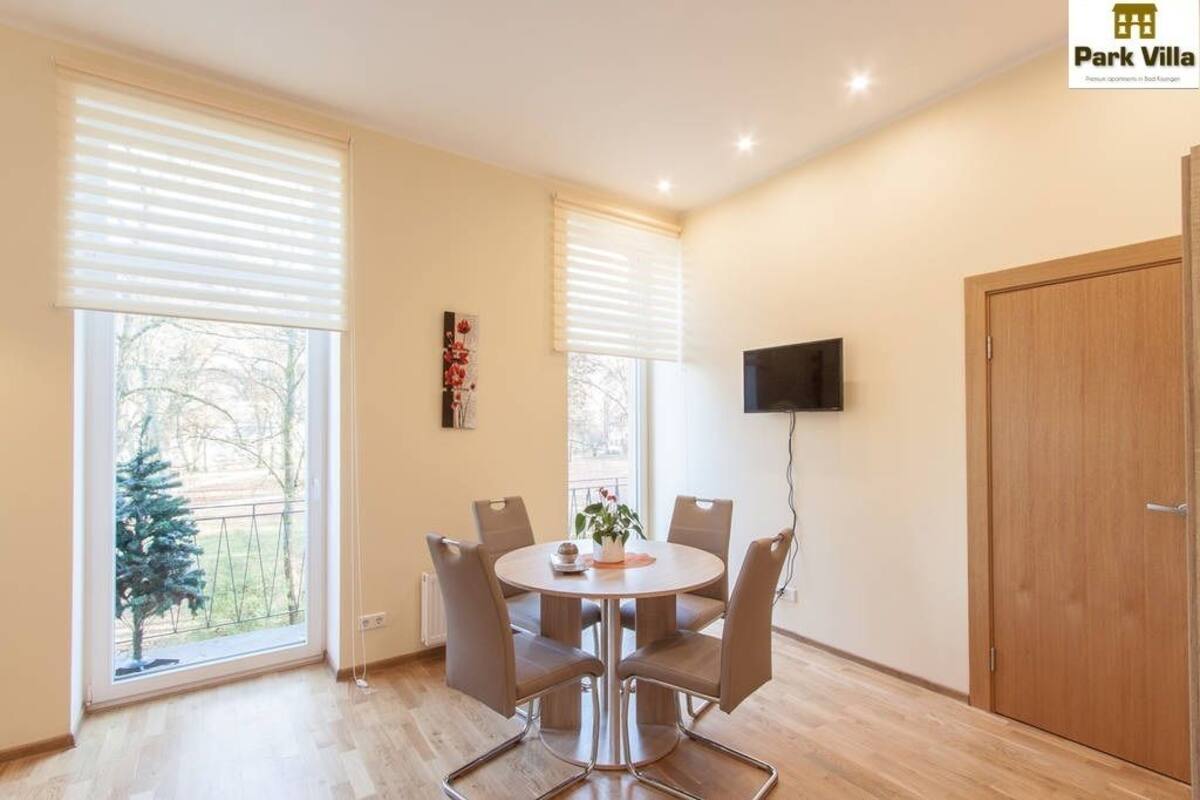 Een eetruimte met een ronde tafel en stoelen, grote ramen met zonwering, een flatscreen televisie aan de wand en een houten deur.