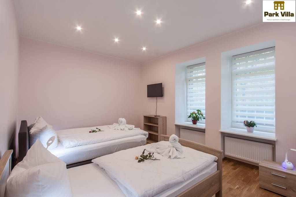 A hotel room with two beds, a TV, a window with blinds, and a small plant on the windowsill.