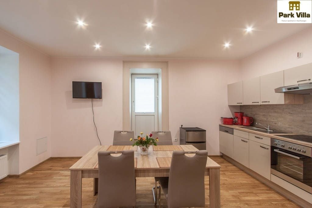 A modern kitchen with a dining area, a flat-screen TV mounted on the wall, and a window with blinds.