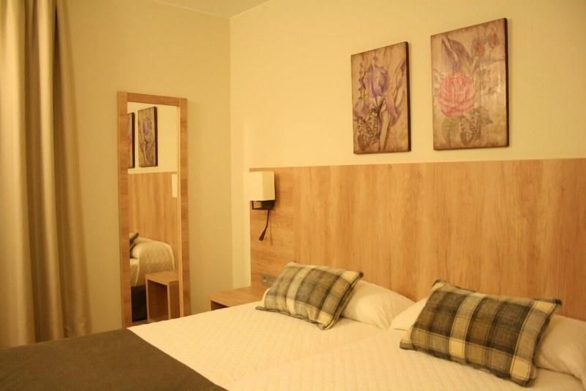 Una habitación de hotel con cabecera de madera, dos camas con almohadas de cuadros, una mesita de noche, una lámpara y dos cuadros con motivos florales en la pared.