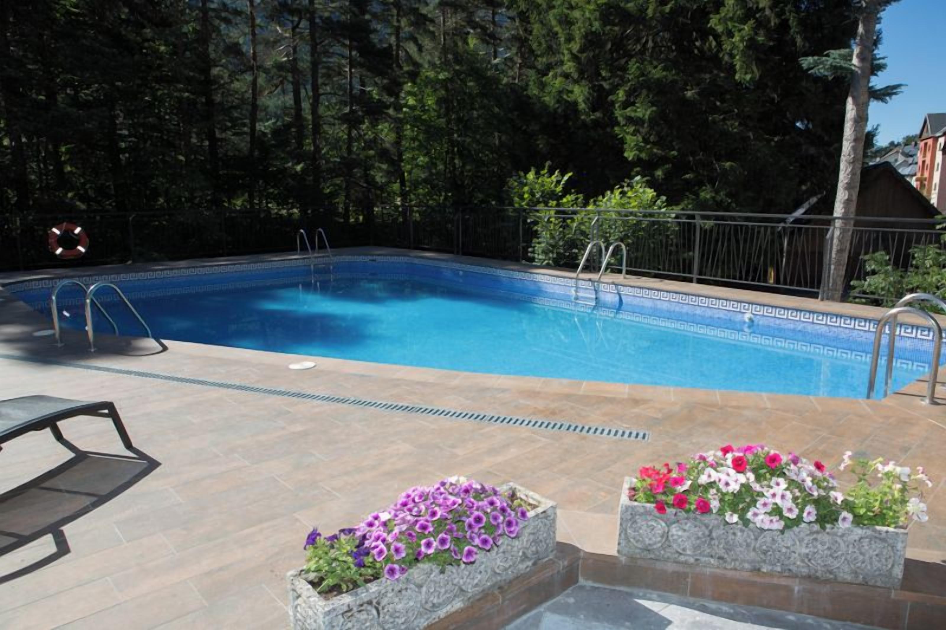 Una piscina con escalones, rodeada por una terraza de madera y macetas de piedra con flores.