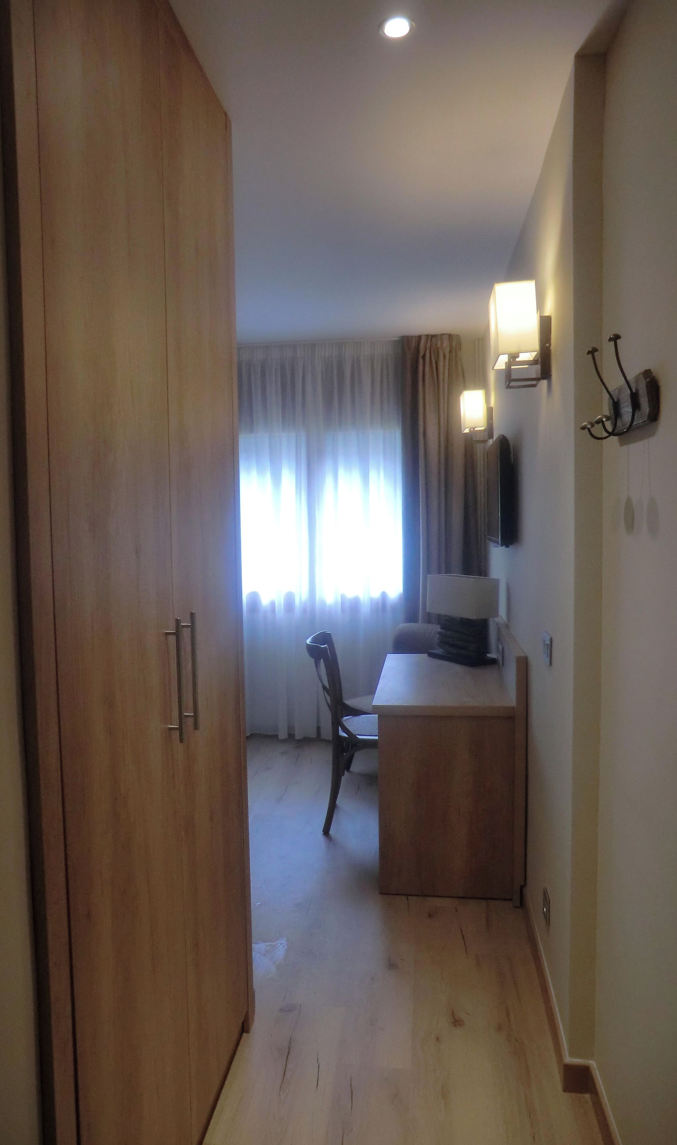 A hotel room with a wooden wardrobe, a desk with a chair, a flat-screen TV, and a window with curtains.