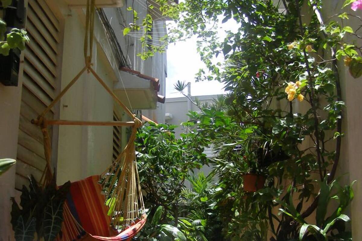 A narrow, sunlit courtyard with a hammock, potted plants, and a tiled floor.