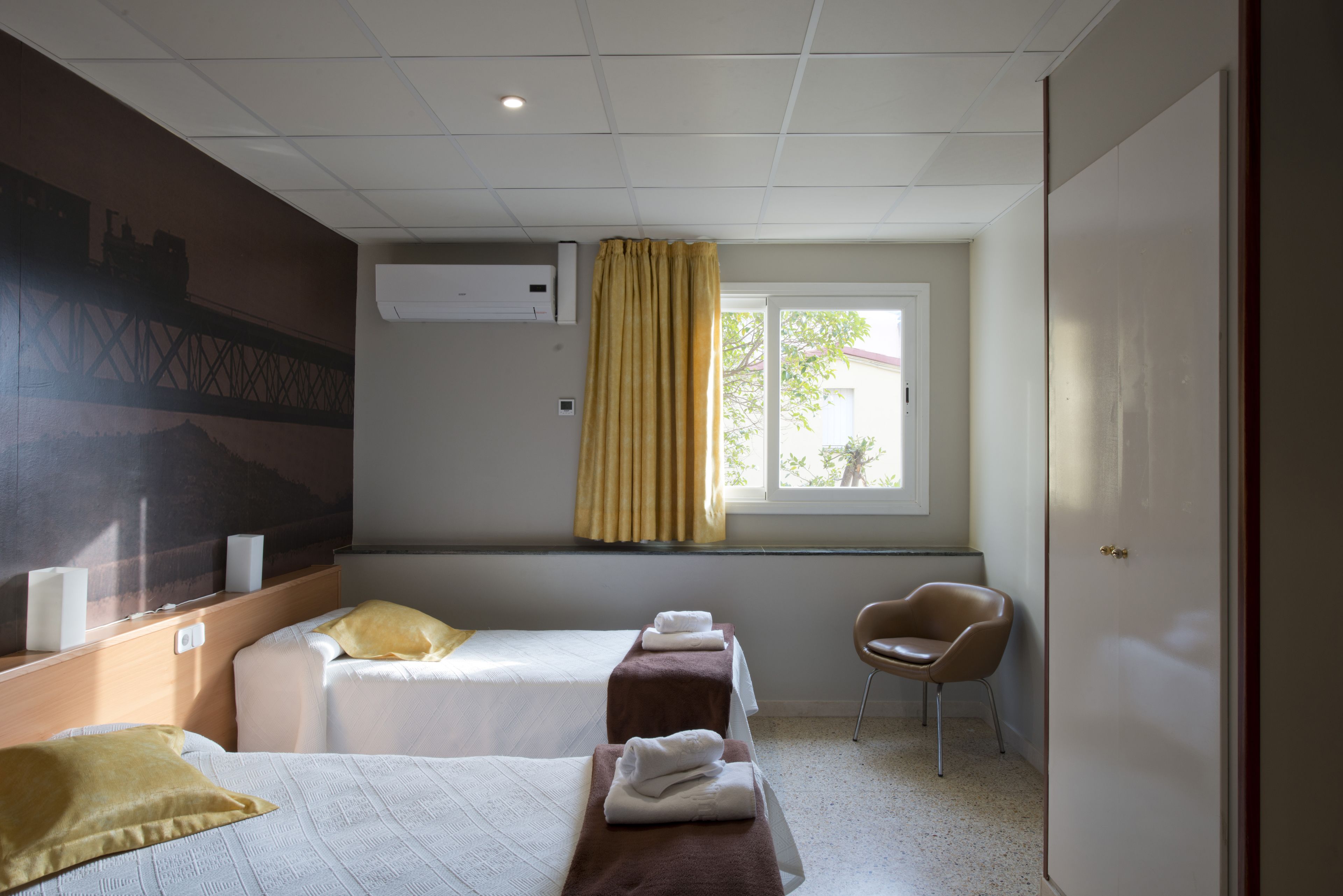 A hotel room with two beds, a window with curtains, a chair, and a wall-mounted air conditioner.