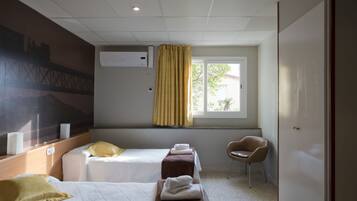 A hotel room with two beds, a window with curtains, a chair, and a wall-mounted air conditioner.