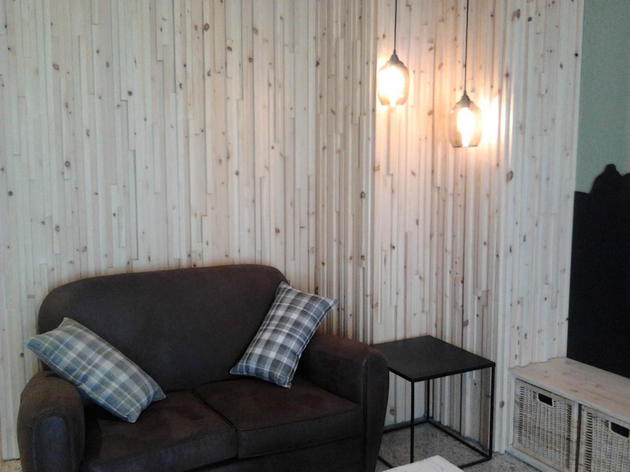 Een kamer met een donkerbruine bank, twee geruite kussens, een klein zwart bijzettafeltje en een witte kast tegen een houten wand met twee hanglampen.