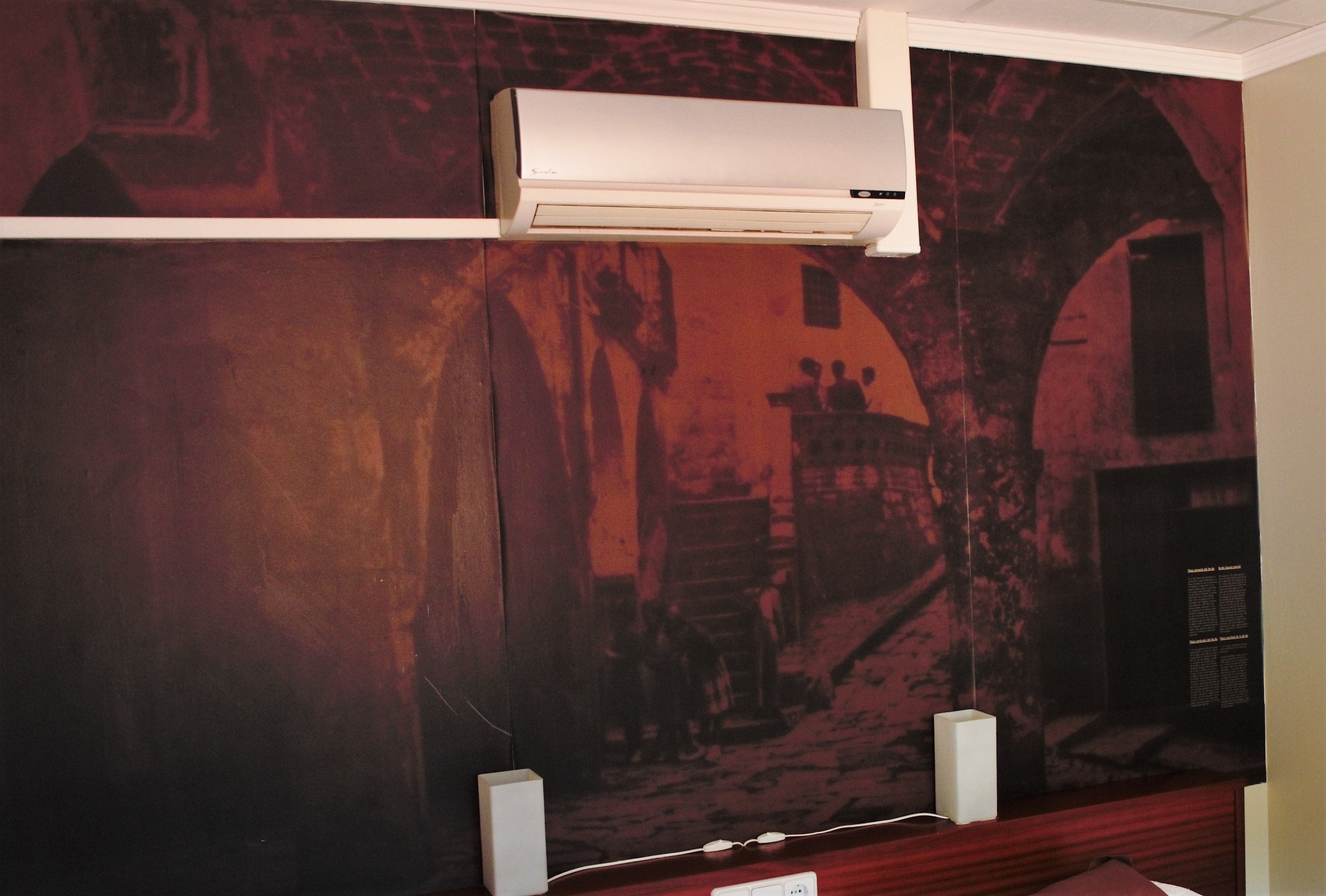 Habitación con un mural que representa una calle de pueblo antiguo, un aire acondicionado blanco y un escritorio de madera con una silla.